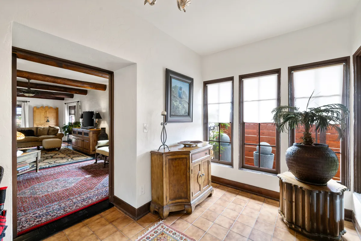 A spacious living area with a wooden cabinet, large windows that flood the room with sunlight, framed artwork, and lush indoor plants. The seating arrangement feels cozy and ideal for socializing or relaxing while enjoying views of the dramatic landscape outside.