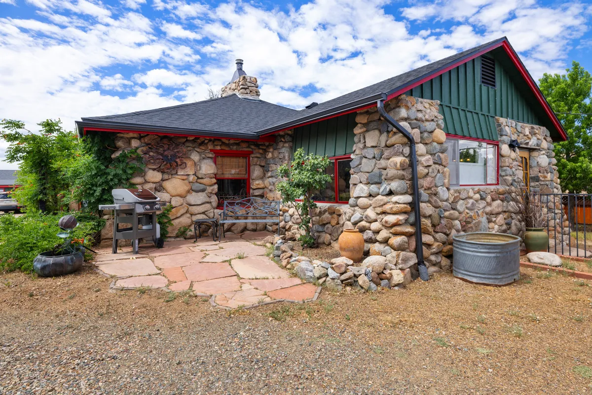 The image showcases a charming stone house with vibrant green accents, set against dramatic Prescott rock formations. The inviting patio area is adorned with decorative pottery, an outdoor grill, and comfortable seating, creating a cozy atmosphere ideal for relaxing or dining outdoors.