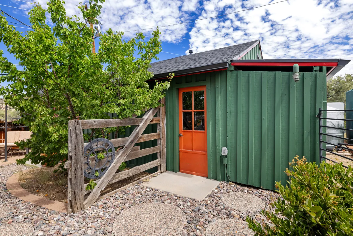 A welcoming exterior shot featuring green wooden siding, an inviting orange door, a wooden fence, a lush tree providing shade, and a pebble walkway. The scene suggests a peaceful entry point and the promise of outdoor relaxation or exploration.