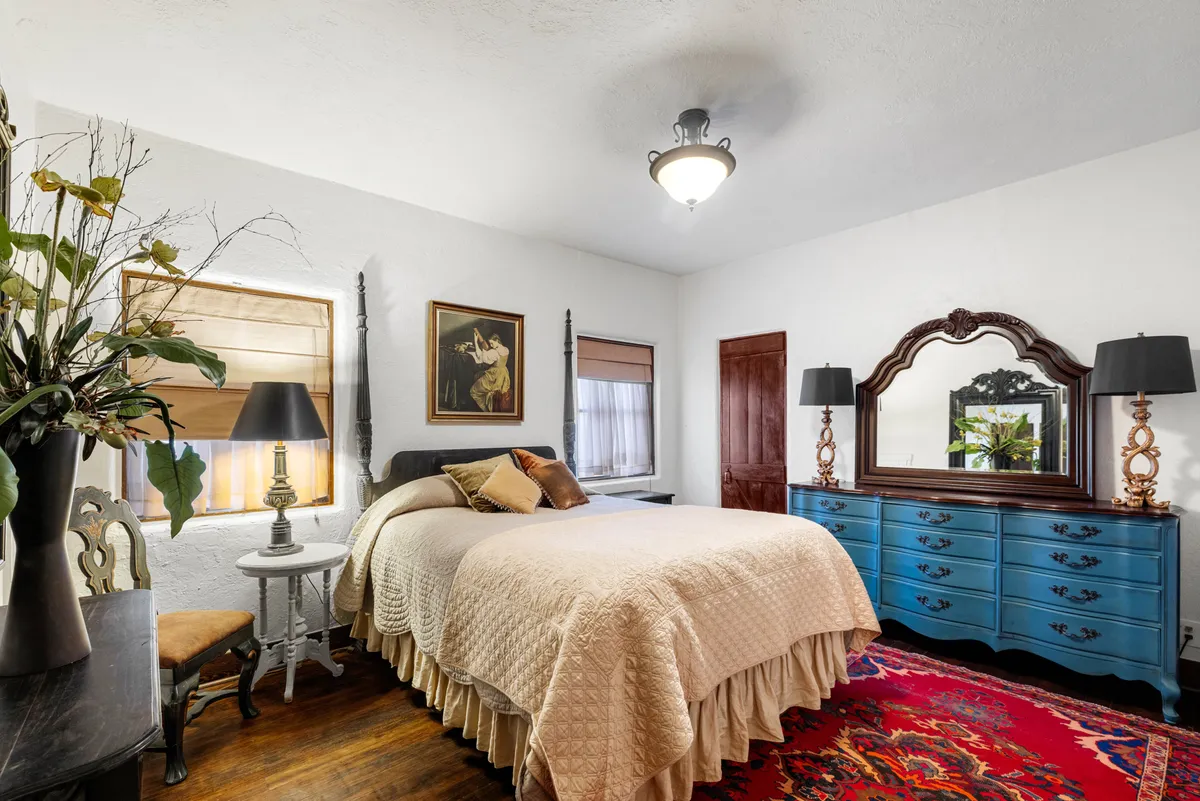 A cozy bedroom featuring a bed with cream bedding and soft linens, a blue dresser, black lamps for ambient lighting, an art piece on the wall, and a colorful rug. Gentle natural light spills in, creating a tranquil and welcoming atmosphere ideal for restful nights.