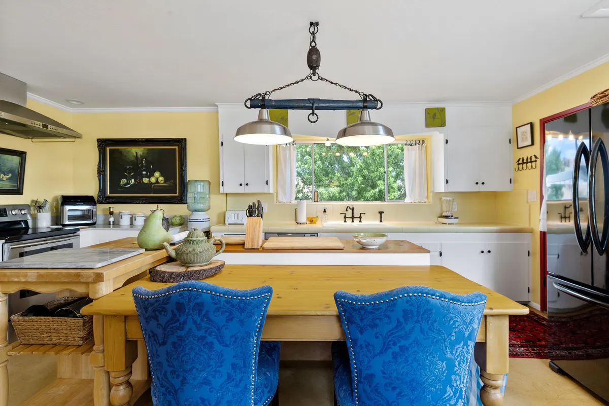 A well-lit dining area featuring a generous wooden table surrounded by blue upholstered chairs. The kitchen is visible in the background, equipped with a kitchen island, a black refrigerator, and a large window streaming in natural light. The setup suggests both a space for meals and an inviting spot for gathering or planning.
