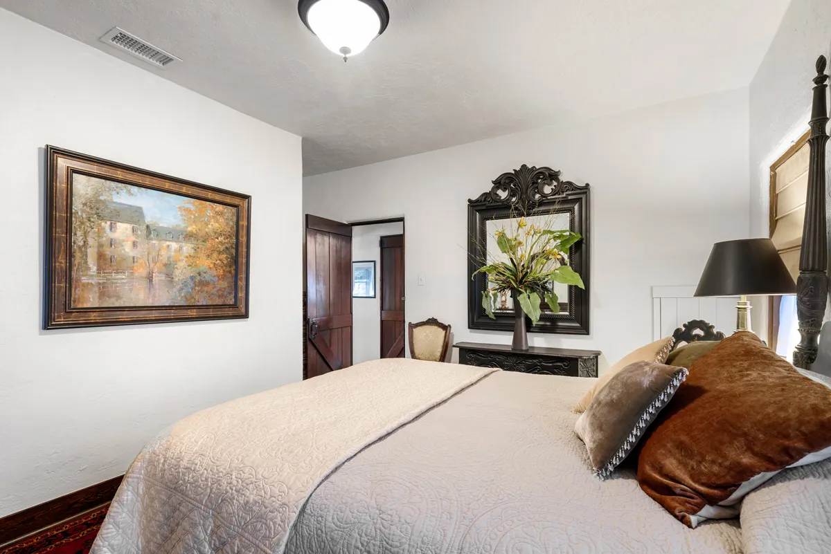 A tranquil bedroom space with a bed adorned with decorative pillows, a framed painting, an ornate mirror with flowers, and a stylish lamp. The arrangement and decor emphasize comfort and peacefulness, reinforcing the idea of a restful retreat.