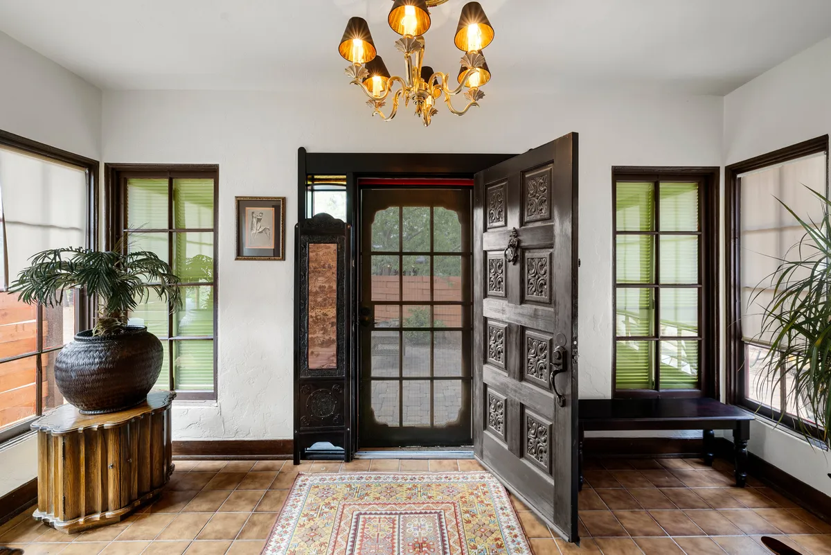 An ornate front door opens into a warmly lit entryway featuring a decorative rug, chandelier, and potted plant. The view emphasizes the welcoming entrance, symbolizing both literal and figurative transitions from the sanctuary of home to the vibrant outside world.