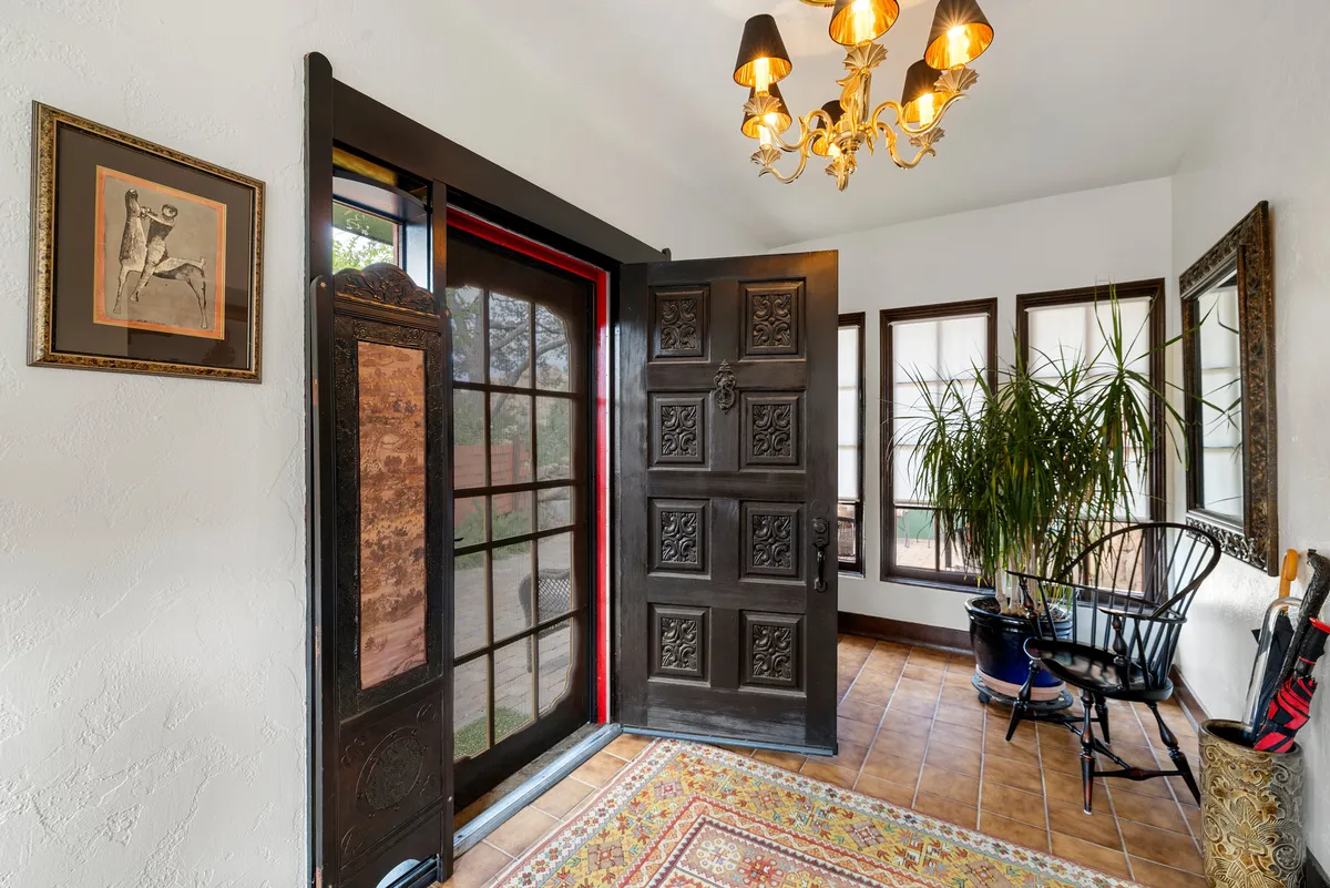 This image captures the entryway with an ornate front door, chandelier, framed artwork, indoor plant, and a decorative rug. There’s a seating area that feels welcoming, filled with soft natural light streaming in, perfect for greeting guests and making a strong first impression.